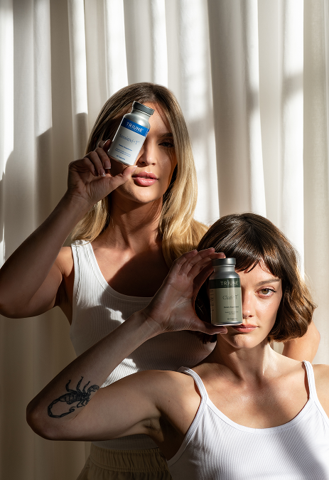 Two women in white tops hold bottles of Triune supplements near their faces